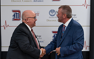 Two men smile and shake hands and look at each other, with logos for University of ɫۺϾþ Mercy and Macomb Community College on a board behind them.