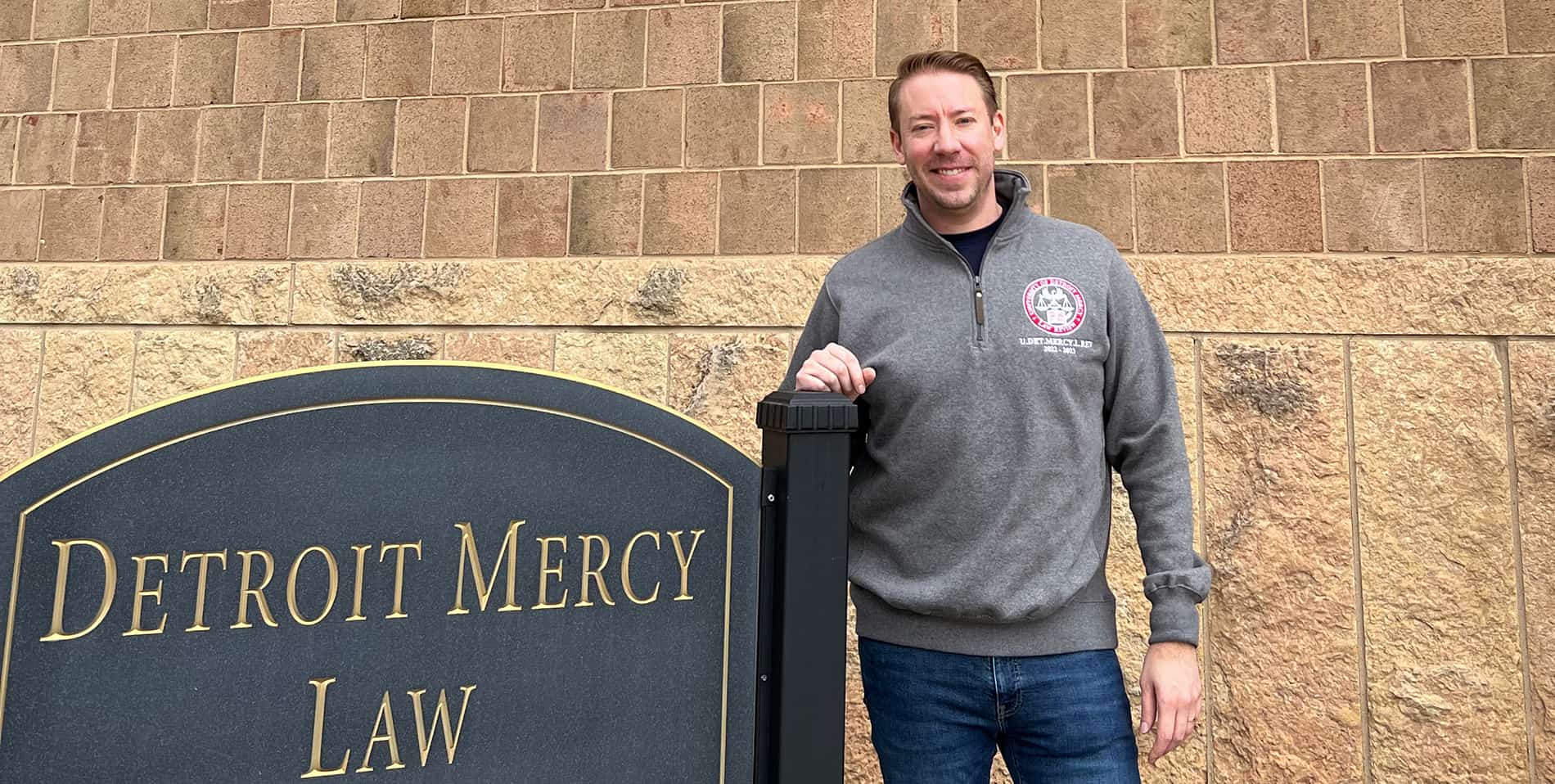 Matthew Snyder stands in front of the School of Law, next to a sign that reads ɫۺϾþ Mercy Law.