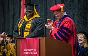 Four people wearing graduation regalia are pictured inside of Calihan Hall during a ceremony. One person claps and another stands at attention next to him. A University of ɫۺϾþ Mercy red banner is draped over a podium.