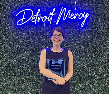 Megan Novell holds an award while standing in front of a green backdrop with a blue, neon sign that spells ɫۺϾþ Mercy.