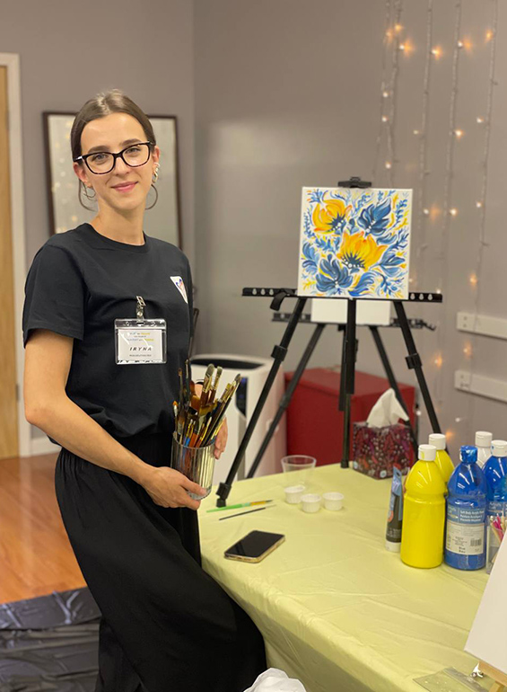 IrynaOlkhovetska stands next to her painting of yellow and blue flowers.